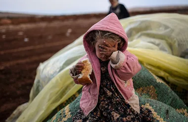 Child in pink hoodie covering face with one hand, holding bread in the other, sitting outdoors on tarps.