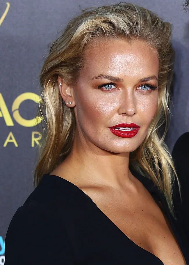 A person with blonde hair and red lipstick poses at an event, wearing a black outfit against a dark background.