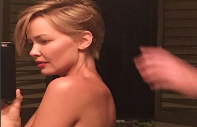 Person taking a selfie in a mirror, with short hair and a bare shoulder.