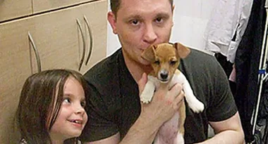 Man holding small puppy with a young girl smiling nearby in a room with wooden cabinets.