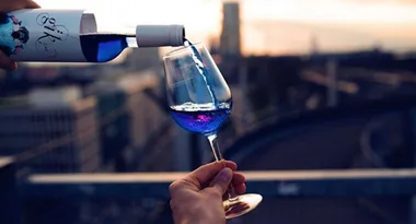 Blue wine being poured into a glass against a blurred cityscape backdrop at sunset.