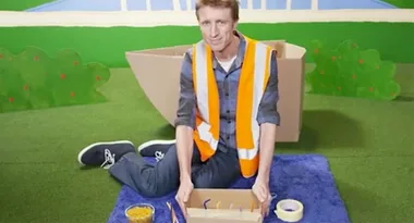 Man in safety vest sitting on a grass mat with a cardboard box, tape, and tools.