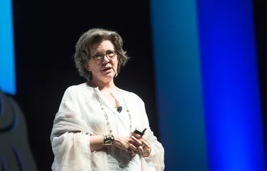 A person stands on stage wearing glasses and a light outfit, speaking into a microphone, with blue lights in the background.