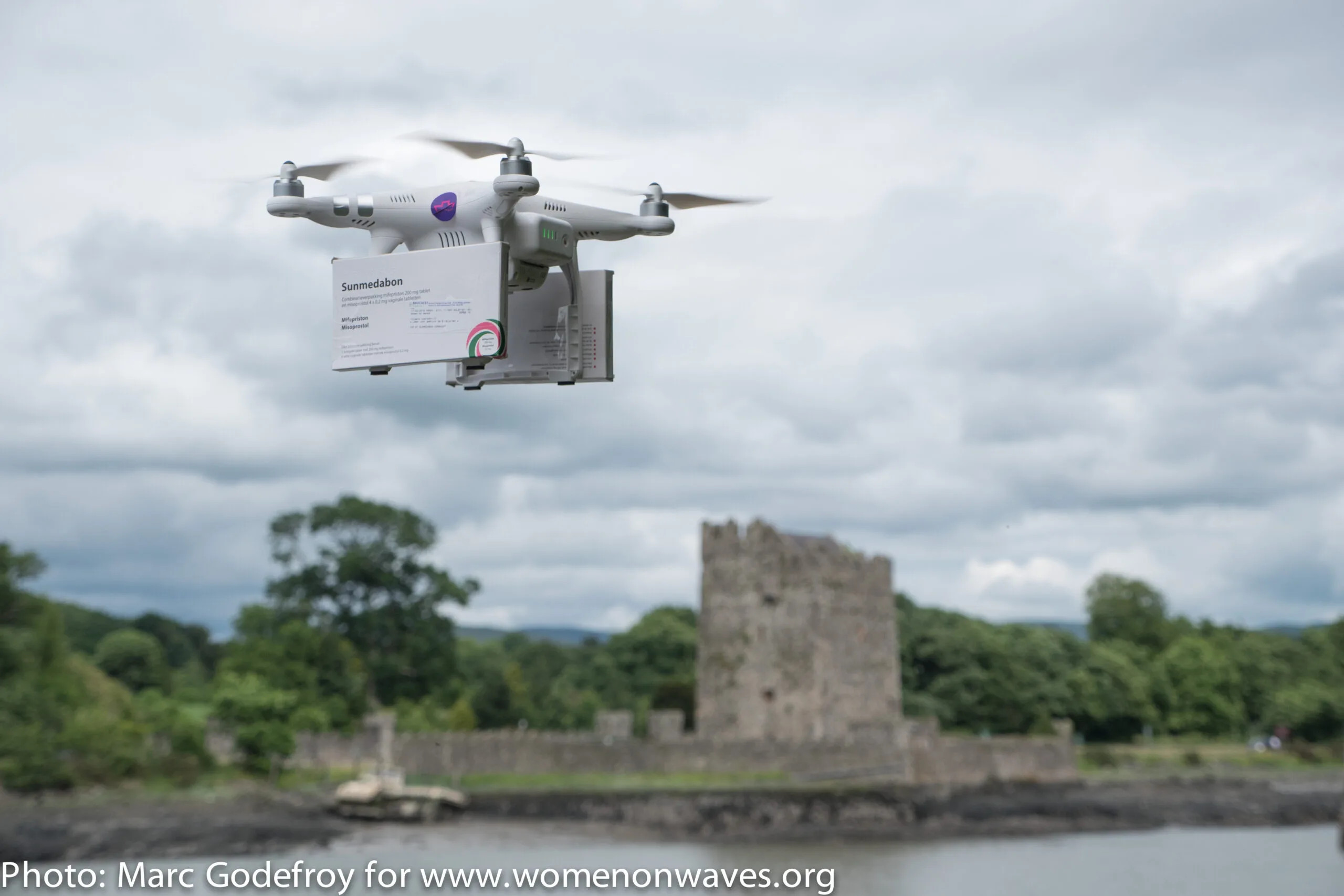 Drone carrying a package, flying over a scenic landscape with an ancient stone structure in the background.