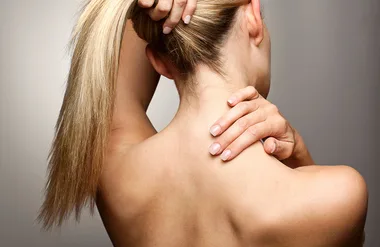 A person with blonde hair seen from the back, holding their neck and shoulder.