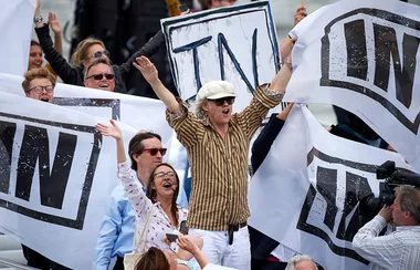 Group of people holding banners with "IN" printed, cheering enthusiastically, one man in striped shirt and hat in the center.