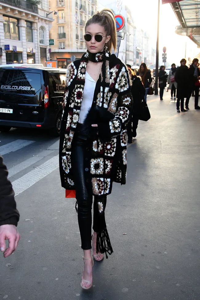 A woman in sunglasses and a crocheted coat walks down a city street, surrounded by people and vehicles.