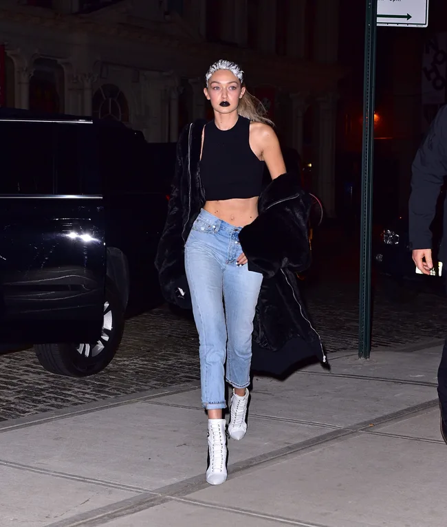 A woman in a black crop top and jeans with dark lipstick walks on a city street at night.