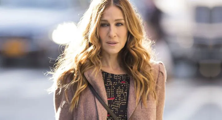 A woman with long wavy hair in a coat, standing on a city street, looking directly at the camera.