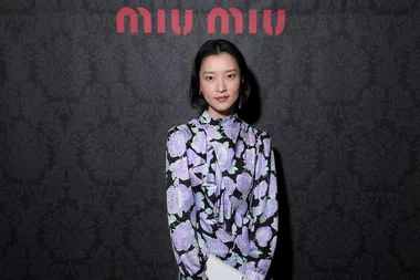 Woman in floral dress posing in front of a patterned black wall with "Miu Miu" logo.