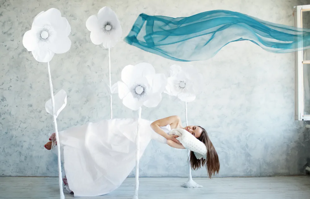 A woman in a white dress levitates among giant paper flowers, holding a pillow, with a blue fabric floating above.