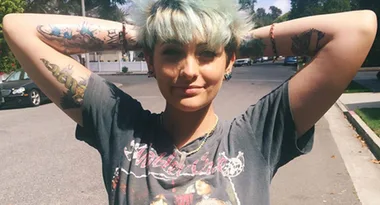 A person with short blue hair and tattoos poses confidently with arms raised, wearing a graphic T-shirt on a sunny street.