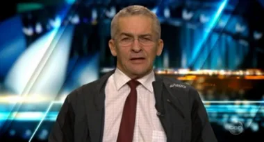 Man in glasses and a formal shirt with a tie, wearing a jacket, against a blurred background of lights and shapes.