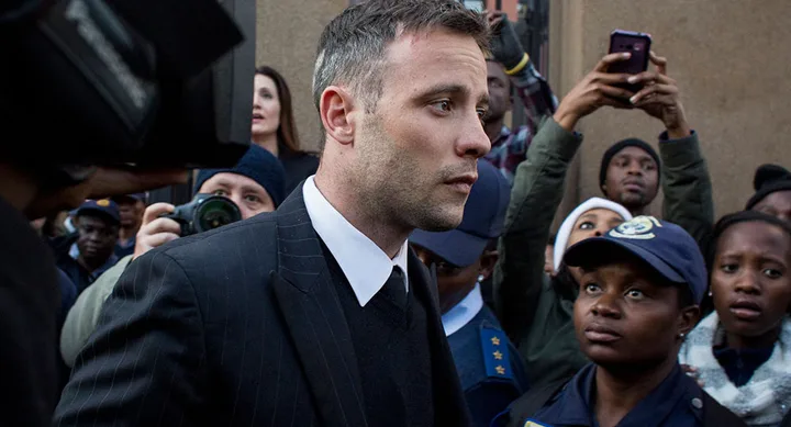 A man in a suit surrounded by police and onlookers outside a building.