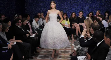 Model walking down the runway in a strapless, textured white dress at a fashion show, surrounded by seated audience.