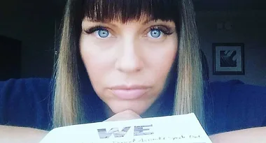 Woman with blue eyes and straight hair, holding a book with "WE" visible on the cover.