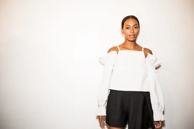 A person in a white off-shoulder top and black shorts stands against a plain background.