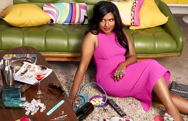 Woman in a pink dress sitting on a shag rug, surrounded by a drink, a magazine, and a colorful couch in the background.