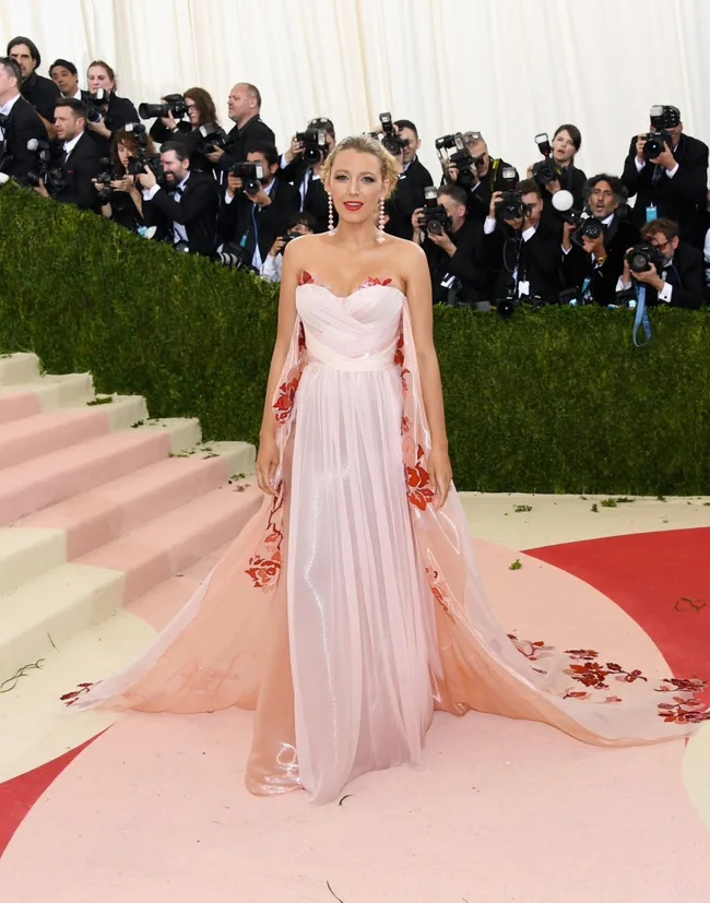A person in an elegant strapless pink dress with floral details on a red carpet, surrounded by photographers.