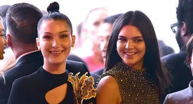 Two women smiling, one with a topknot hairstyle wearing a black outfit, and the other with long hair in a beaded dress.