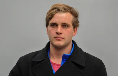 A man with short blonde hair wearing a black coat and blue shirt stands against a plain gray background.
