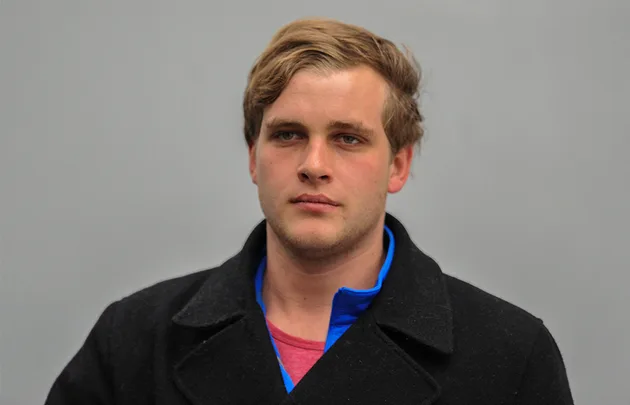 A man with short blonde hair wearing a black coat and blue shirt stands against a plain gray background.