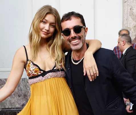 Two individuals posing together; one wears a yellow dress, and the other wears sunglasses and a dark jacket.