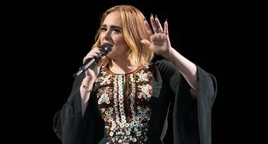 A person singing passionately on stage, wearing a black dress with sequins, holding a microphone.