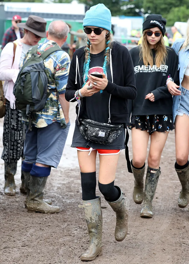 Festival-goers walking in muddy field, wearing wellies and casual clothing, with one person using a smartphone.