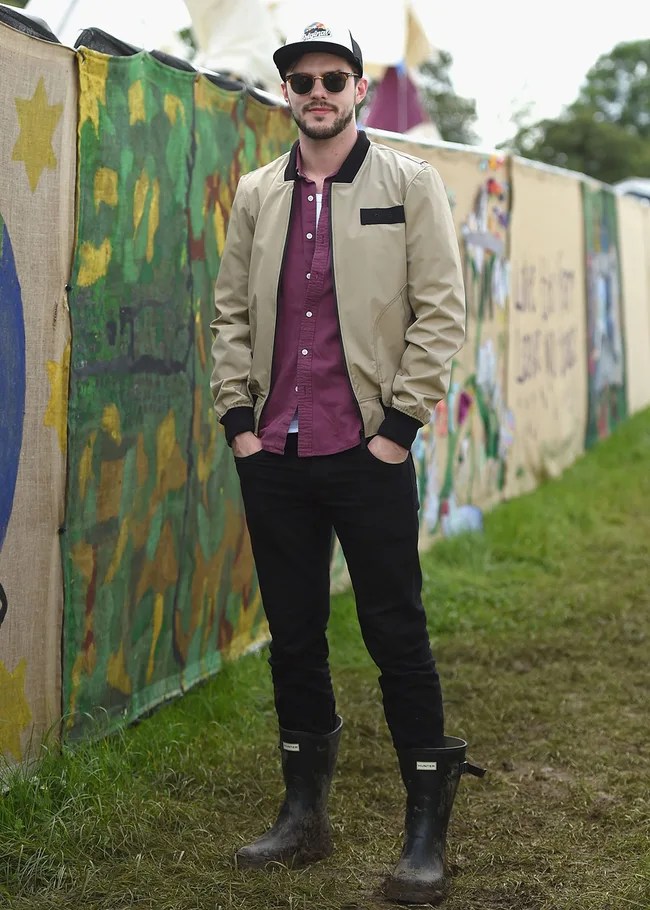 Man in a beige jacket and cap, wearing muddy boots, stands outdoors near vibrant painted panels.