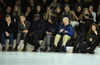 People seated in front row at a fashion show, some holding cameras, all focused on the runway.