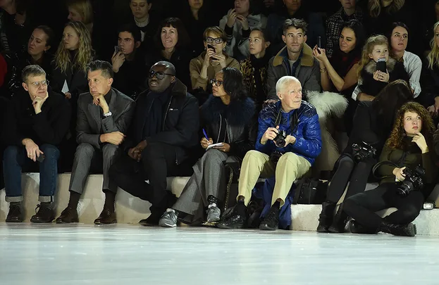 People seated in front row at a fashion show, some holding cameras, all focused on the runway.