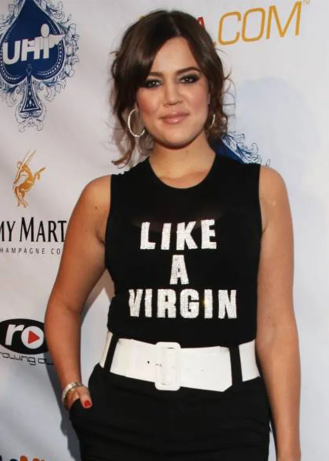 Woman in black sleeveless top with "Like a Virgin" text, large white belt; step-and-repeat background.