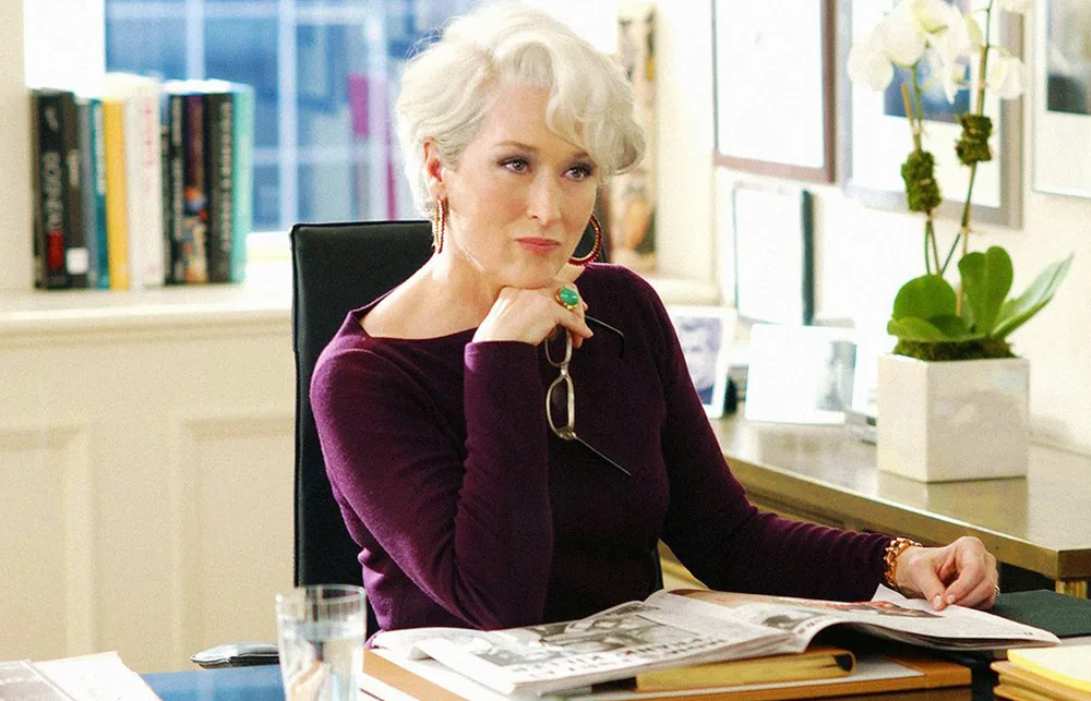 A woman with silver hair sits at a desk, holding glasses, with a thoughtful expression in a stylish office.