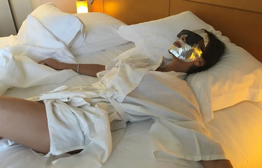 Person relaxing on a bed wearing a white robe, sunglasses, and a metallic face mask.