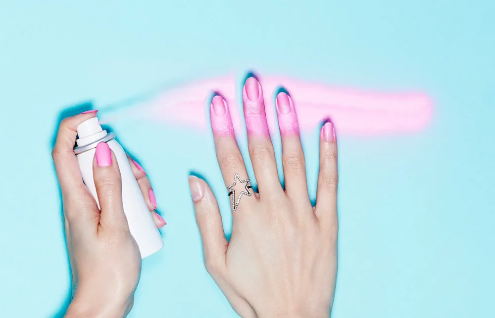 Spray painting nails pink on a hand with a star ring, against a light blue background.