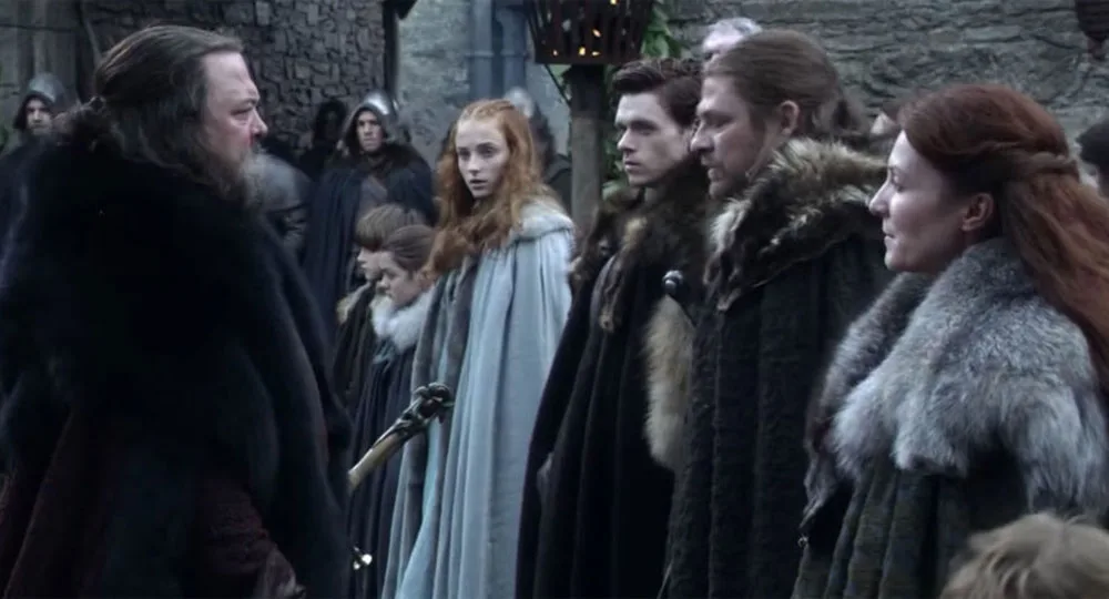 A group of people in medieval clothing stand outdoors, facing an older man in dark fur.