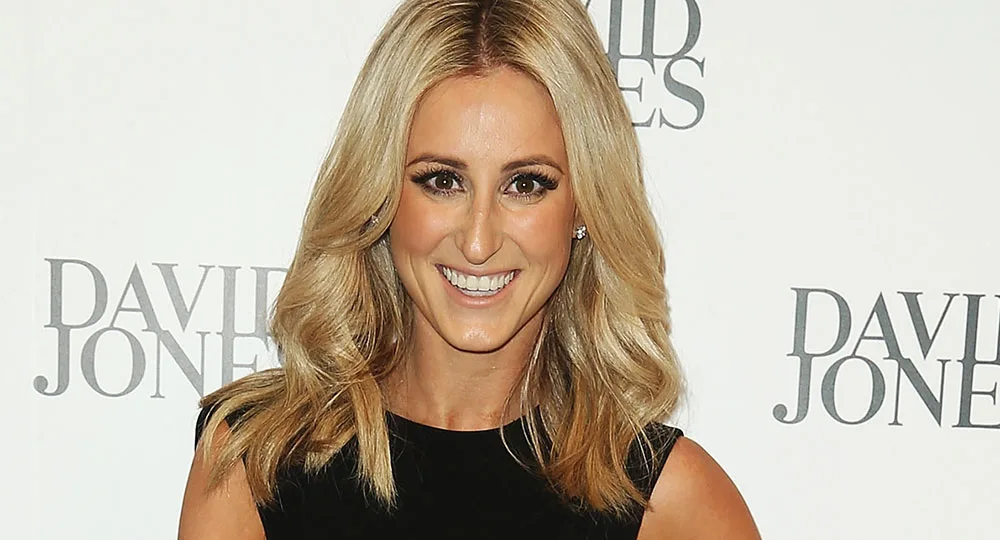 A woman with long blonde hair in a black dress smiles at a David Jones event backdrop.