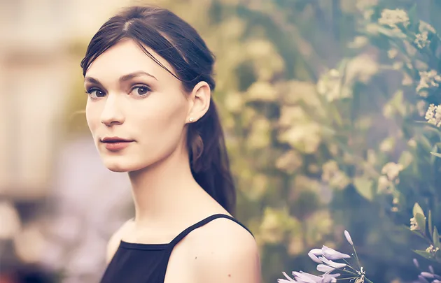A woman with dark hair in a black dress stands against a blurred floral background.