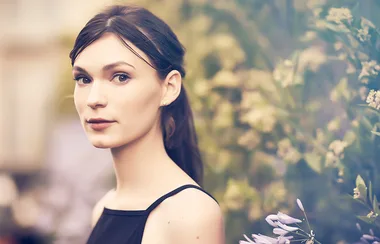 A woman with dark hair in a black dress stands against a blurred floral background.