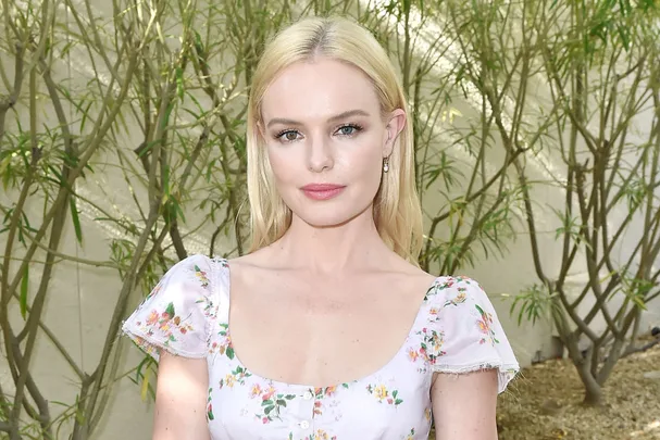 A woman with blonde hair in a floral dress posing in front of green foliage.