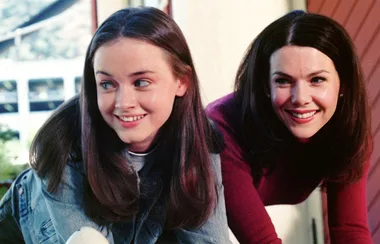 Two women smiling together, one younger with long brown hair in a denim jacket, the other older in a red sweater.