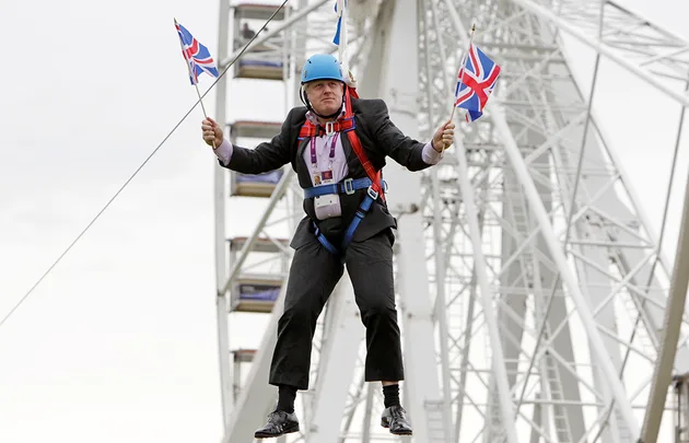 Man in a suit on a zip line holds two Union Jack flags, background features a Ferris wheel structure.