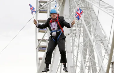 Man in a suit on a zip line holds two Union Jack flags, background features a Ferris wheel structure.