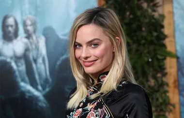 A person with long blonde hair, wearing a black embroidered outfit, smiles at an event with a blurred poster in the background.