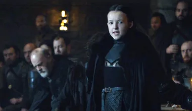 A young girl in medieval clothing speaks confidently in a dimly lit room with men observing her intently.