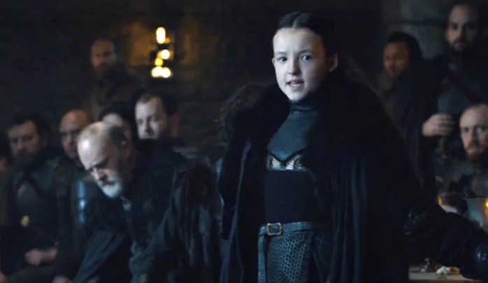A young girl in medieval clothing speaks confidently in a dimly lit room with men observing her intently.