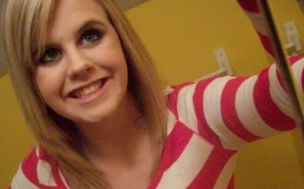 A young woman with blonde hair wearing a red and white striped top, smiling in a selfie against a yellow background.