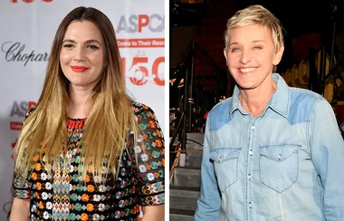 Two women smiling; one in colorful patterned dress, the other in a denim shirt, indoors with a backdrop and stage setting.