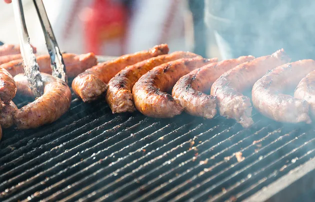 Grilled sausages cooking on a barbecue with tongs turning them, smoke rising from the grill.